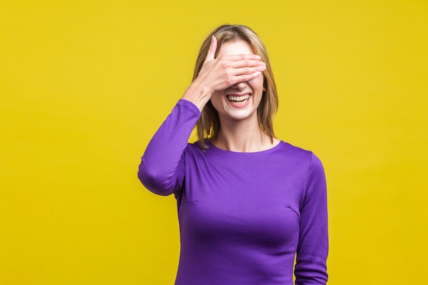 Porträt einer schüchternen, positiven Frau in einem eleganten lila Kleid, das die Augen mit der Hand bedeckt und schüchtern lacht, um eine Studioaufnahme im Innenbereich isoliert auf gelbem Hintergrund zu sehen