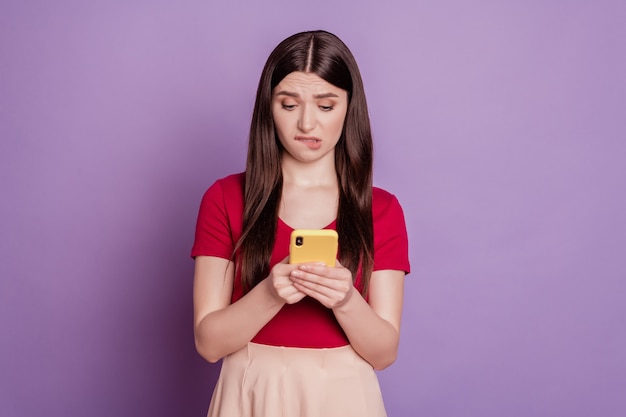 Porträt einer schuldigen, traurigen mädchenhaften Dame, die Telefon-Look-Bildschirm-Bisslippe auf violettem Hintergrund hält