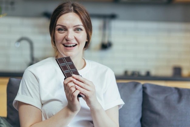 Porträt einer schwangeren Frau, die eine Tafel mit köstlichen Schokoladenbonbons isst. Sitzen auf der Couch zu Hause in Hauskleidung, die lächelnd in die Kamera schaut. Zufrieden liebt Süßigkeiten