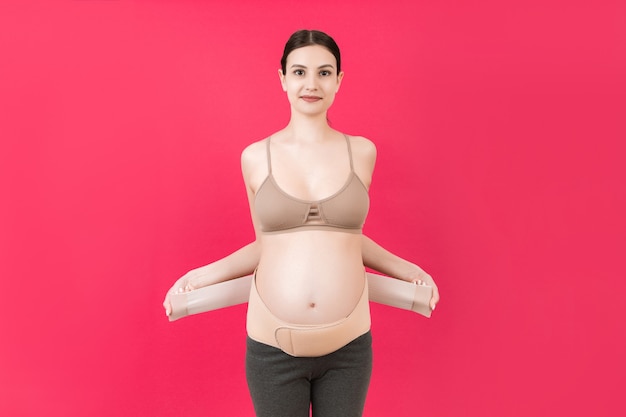 Porträt einer schwangeren Frau, die einen Stützverband anlegt, um Rückenschmerzen auf rosafarbenem Hintergrund mit Kopienraum zu reduzieren. Orthopädisches Bauchstützgurtkonzept.