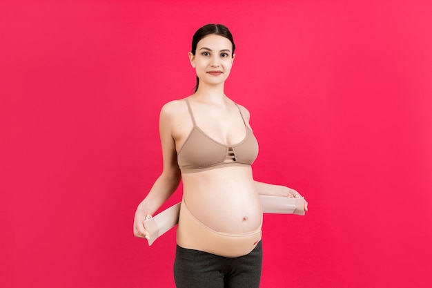 Porträt einer schwangeren Frau, die einen Stützverband anlegt, um Rückenschmerzen auf rosafarbenem Hintergrund mit Kopienraum zu reduzieren. Orthopädisches Bauchstützgurtkonzept.