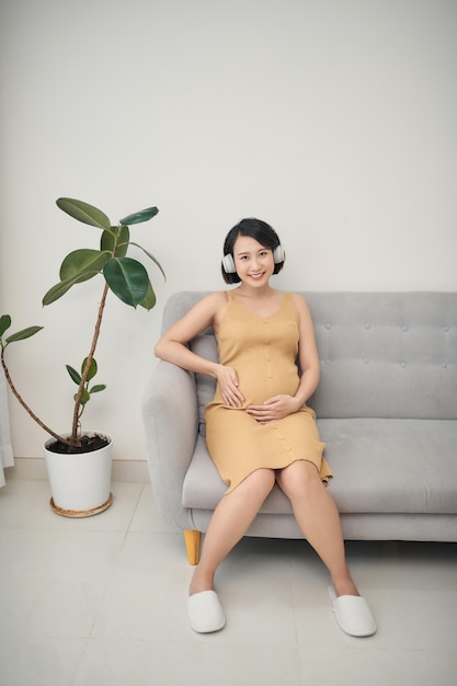 Porträt einer schwangeren Frau, die zu Hause auf dem Sofa sitzt und Musik über Kopfhörer hört