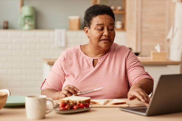 Porträt einer schwarzen Seniorin, die einen Laptop benutzt, während sie das Frühstück in der gemütlichen Küche zu Hause genießt