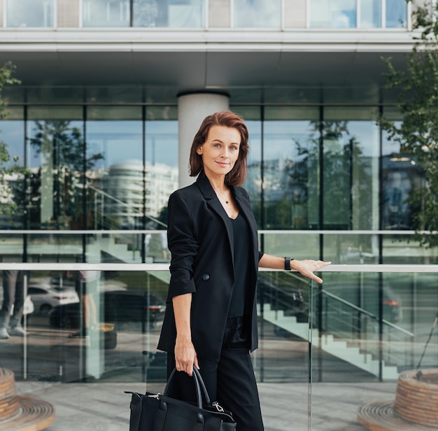 Porträt einer selbstbewussten Frau mittleren Alters in formeller Kleidung mit einer Tasche gegen ein Bürogebäude
