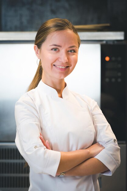 Porträt einer selbstbewussten und lächelnden jungen Köchin in weißer Uniform, professionelle Küche im Hintergrund