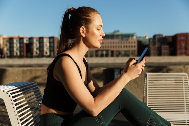 Porträt einer sonnenbeschienenen sportlichen Frau mit Trainingsanzug, die Smartphone hält und auf einem Stuhl sitzt, während sie in der Stadtstraße trainiert?