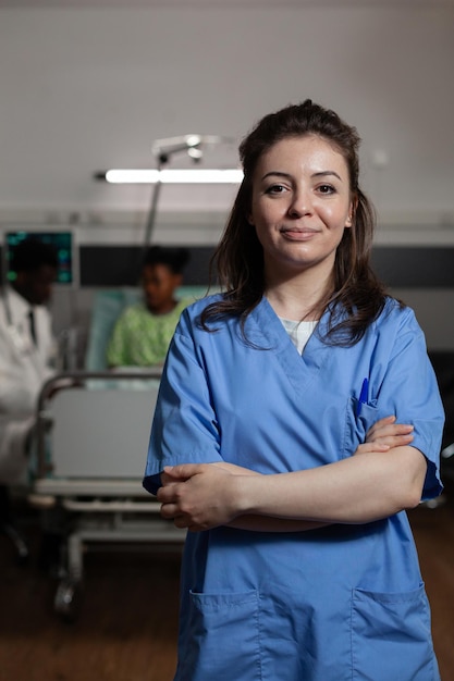 Porträt einer spezialisierten Therapeutenkrankenschwester, die während eines Arzttermins auf der Krankenstation steht. Im Hintergrund berät der Arzt einen kranken Patienten und bespricht die medizinische Behandlung. Medizinische Dienstleistungen