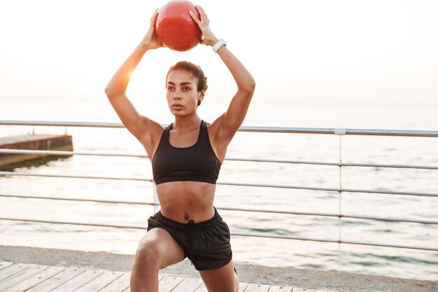 Porträt einer sportlichen Frau in sportlicher Kleidung, die mit Fitnessball hockt, während sie morgens am Meer trainiert?