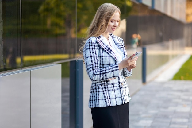 Porträt einer stilvollen, attraktiven Geschäftsfrau mit blonden Haaren in karierter Jacke und Rock mit Smartphone im Freien, die glücklich aussieht, während sie eine gute Nachricht liest, die in der Nähe eines Bürogebäudes steht