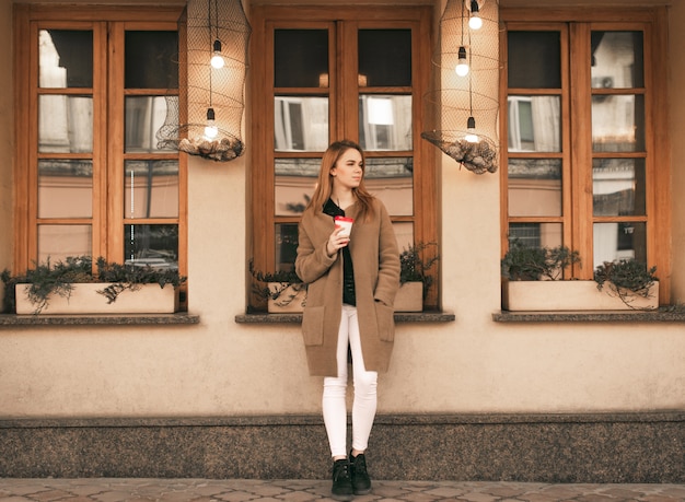 Porträt einer stilvollen Dame in voller Länge, die mit einer Tasse Kaffee in den Händen steht, einen Mantel vor dem Hintergrund der Wand des Cafés trägt und zur Seite schaut