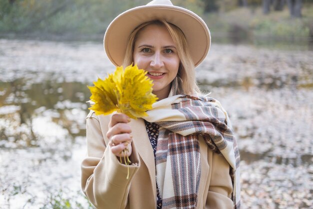 Porträt einer stilvollen Frau in beigem Mantel und Hut hält Herbstblätter im Herbstpark im Herbst
