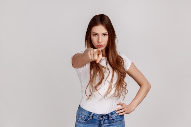 Porträt einer strengen unzufriedenen jungen Frau, die mit dem Zeigefinger eine Wahl trifft und herrisch und unzufrieden aussieht und schuldiges Tragen von weißem T-Shirt zeigt Studioaufnahme im Innenbereich isoliert auf grauem Hintergrund