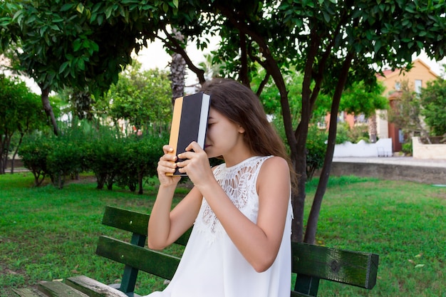 Porträt einer Studentin, die vom Lernen müde ist, sitzt draußen auf einer Bank und hält ein Buch.