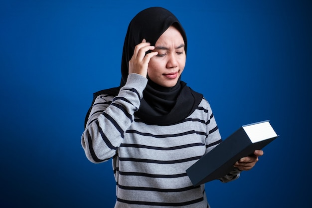 Porträt einer süßen asiatischen muslimischen Frau, die krank und müde ist und zu viel Bücher auf blauem Hintergrund liest