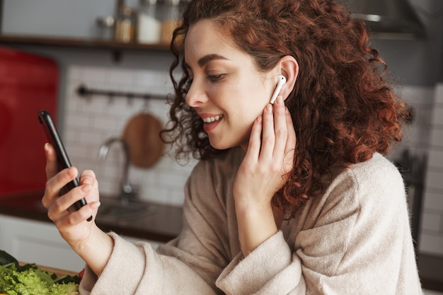 Porträt einer süßen europäischen Frau mit Ohrstöpseln, die Musik auf dem Handy hört, während sie in der Küche zu Hause kocht