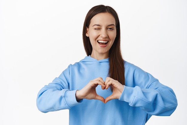 Porträt einer süßen Frau zwinkert, drückt Sympathie aus, zeigt Herzgeste und lächelt kokett, flirtet, steht in blauem Hoodie gegen weiße Wand