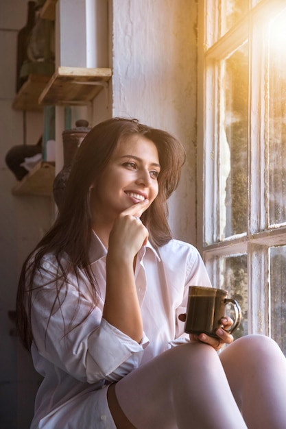Porträt einer süßen jungen Frau in weißem Hemd, die Tee trinkt und durch das Fenster schaut, während sie drinnen auf der Fensterbank sitzt