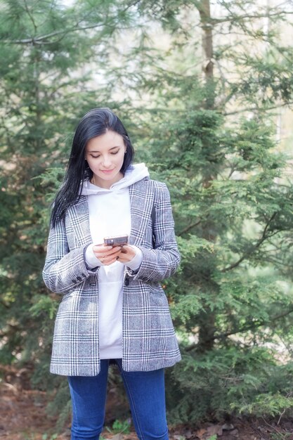 Porträt einer süßen jungen, glücklichen, brünetten hübschen Frau in einem karierten Mantel, die am schönen Frühlingstag mit Handy im Park spazieren geht