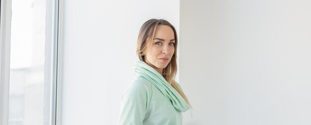 Porträt einer süßen positiven Frau im Minz-Hoodie, die in der Nähe des Fensters steht und sich im Wohnzimmer in ihrem Haus entspannt. Kopieren Sie Platz und Platz für Werbung.