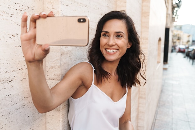 Porträt einer süßen, zufriedenen Frau, die lächelt und ein Selfie-Foto auf dem Smartphone macht, während sie über der Mauer auf der Stadtstraße steht