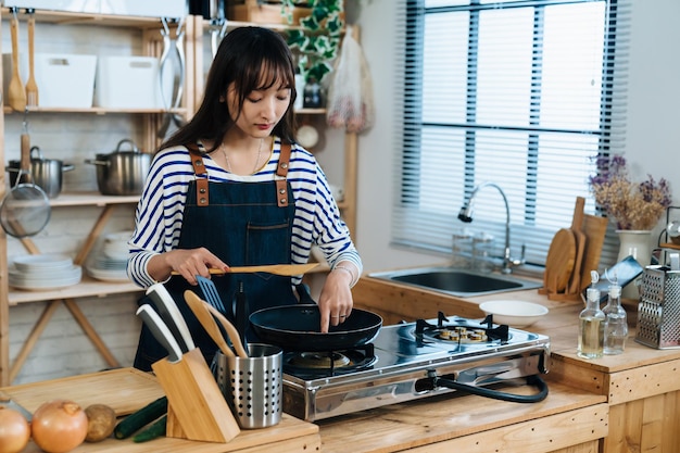 Porträt einer taiwanesischen Hausfrau, die mit dem Finger Essen in der Bratpfanne berührt, um den Geschmack zu schmecken, während sie am Gasherd in einer hellen Küche zu Hause Essen zubereitet