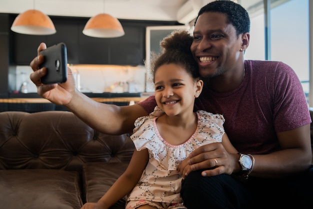 Porträt einer Tochter und eines Vaters, die Spaß zusammen haben und ein Selfie mit Handy zu Hause machen. Monoparentales Konzept.