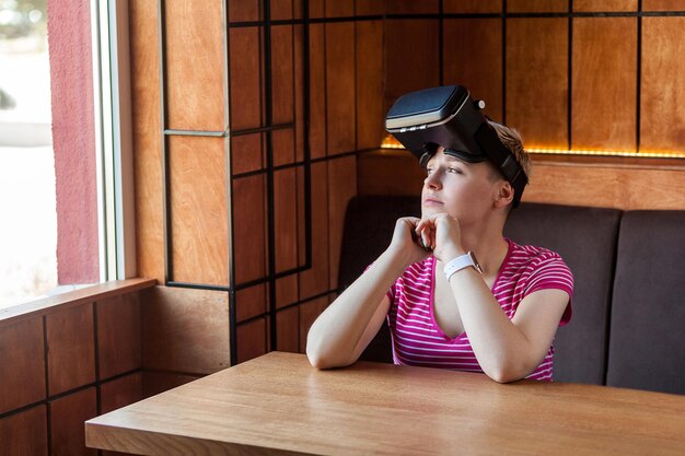 Porträt einer träumenden jungen Frau in rosafarbenem T-Shirt, die im Café sitzt und ein Virtual-Reality-Headset auf dem Kopf trägt und mit denkendem Gesicht auf das Fenster blickt Indoor-Lebensstil