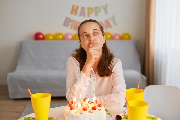 Porträt einer traurigen einsamen jungen erwachsenen nachdenklichen frau, die allein am geburtstagstisch mit kuchen sitzt, das kinn hält und wegschaut, sich nachdenklich ausdrückt und über geburtstagswünsche nachdenkt.