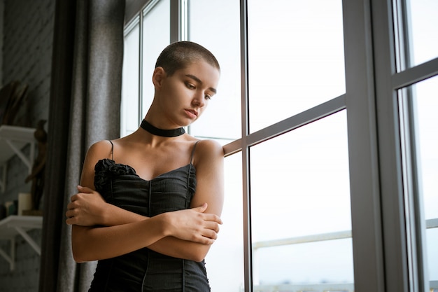 Porträt einer traurigen jungen Frau mit kurzen Haaren, die aus dem Fenster in einem schwarzen Kleid schaut.