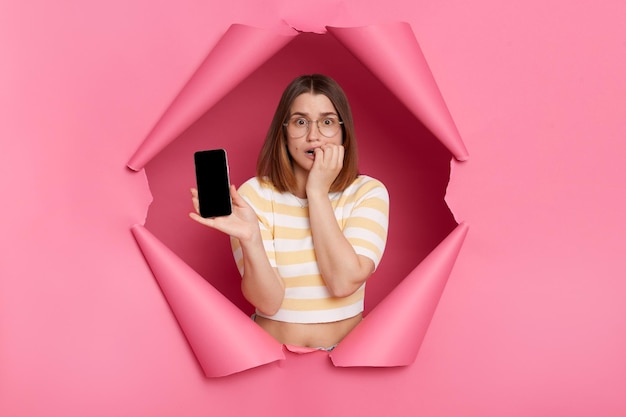Porträt einer traurigen, verärgerten kaukasischen Frau, die durch den rosafarbenen Papierhintergrund bricht und ein Handy mit leerem Display mit Werbefläche für Werbetext zeigt
