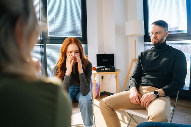 Porträt einer traurigen, weinenden jungen Frau, die Probleme hat, während der Gruppentherapiesitzung im Kreis zu sitzen. Depressive Frauen, die von psychischen Problemen erzählen, bedecken das Gesicht mit den Händen.