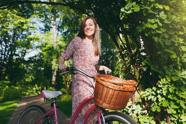 Porträt einer trendigen jungen Frau in langem rosa Blumenkleid, die unter Eichenbogen auf dem Fahrrad mit Korb für Einkäufe, Essen oder Blumen im Freien reitet, süße weibliche Erholungszeit im Frühling oder Sommerpark