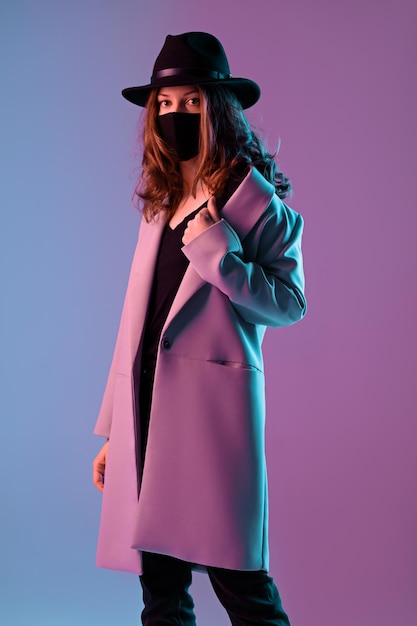Porträt einer trendigen jungen Frau in Mantel, Hut und Schutzmaske in violetter Neonbeleuchtung