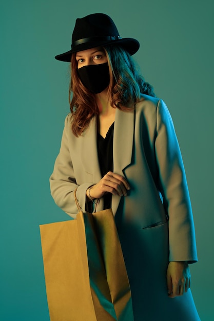 Porträt einer trendigen jungen Frau in Mantel, Hut und Schutzmaske mit Einkaufstüte aus Papier