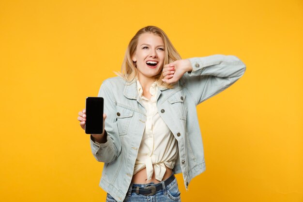 Porträt einer überglücklichen jungen Frau in Denim-Freizeitkleidung hält ein Mobiltelefon mit leerem, leerem Bildschirm isoliert auf gelb-orangefarbenem Wandhintergrund im Studio. People-Lifestyle-Konzept. Mock-up-Kopienbereich.