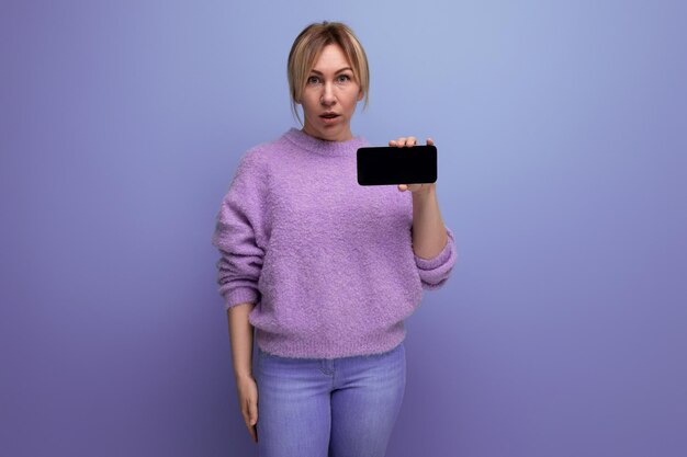 Porträt einer überraschten blonden jungen Frau in einem lila Pullover mit einem Smartphone-Modell horizontal