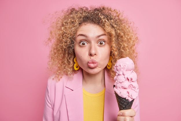 Porträt einer überraschten Frau mit lockigen, blonden Haaren, die in eleganten Kleiderohren gekleidet sind, hält ein großes leckeres Eis und hat einen leckeren Snack während der Sommerzeit, die gegen die rosa Wand posiert