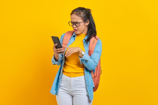 Porträt einer überraschten jungen asiatischen Studentin in lässiger Kleidung mit Rucksack, die Textnachrichten tippt oder über ein Smartphone auf gelbem Hintergrund scrollt, um Feeds in sozialen Netzwerken zu scrollen