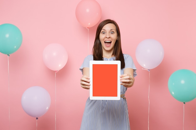 Porträt einer überraschten schockierten jungen glücklichen Frau im blauen Kleid, die Tablet-PC-Computer mit leerem leerem Bildschirm auf pastellrosa Hintergrund mit bunten Luftballons hält. Geburtstagsfeier Konzept.