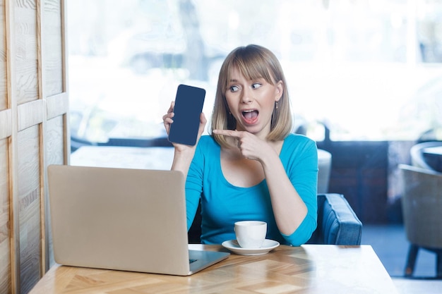 Porträt einer überraschten schönen jungen blonden Frau im blauen T-Shirt, die mit Laptop sitzt, die mobile Anzeige hält und zeigt und mit Erstaunen schaut. Innen, Studioaufnahme.