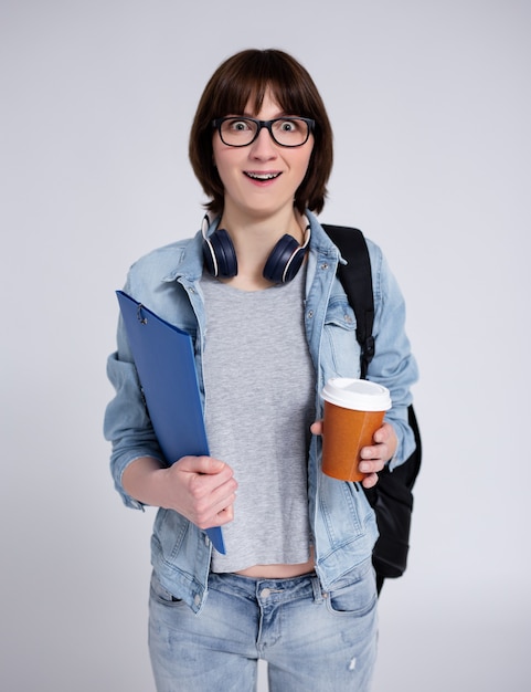 Porträt einer überraschten Studentin in Brillen mit Rucksack, Ordner und Tasse Kaffee auf grauem Hintergrund