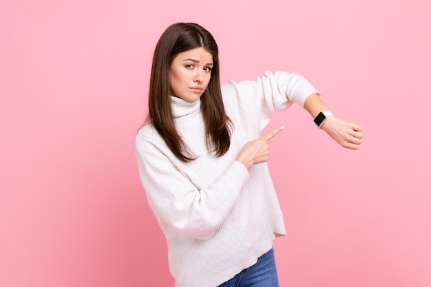 Porträt einer ungeduldigen Frau, die auf eine Armbanduhr zeigt und unzufrieden aussieht und eine Uhr zeigt, um sich mit einem weißen, lässigen Pullover zu beeilen Indoor-Studioaufnahme isoliert auf rosa Hintergrund