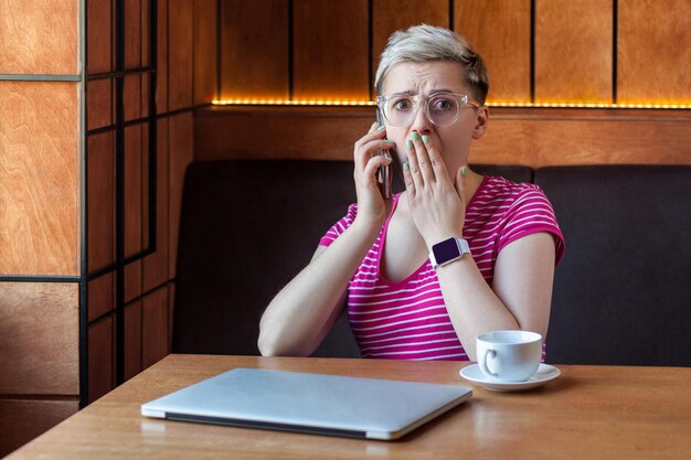 Foto porträt einer unglaublich schockierten jungen geschäftsfrau mit kurzen haaren in rosafarbenem t-shirt und brille sitzt im café, telefoniert und bedeckt den mund und blickt in die kamera