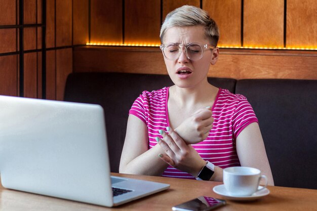 Porträt einer unglücklichen jungen Geschäftsfrau mit kurzen blonden Haaren in rosafarbenem T-Shirt und Brille, die im Café sitzt und den Arm hält und versucht, den Puls mit verzerrtem Gesicht zu fühlen. Indoor, Gesundheitswesen