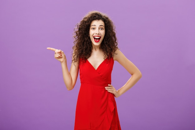 Porträt einer unterhaltenen und amüsierten, fröhlichen europäischen Frau mit lockiger Frisur in rotem Abendkleid, die vor Belustigung und Freude lacht, die nach links zeigt und über lila Hintergrund posiert.