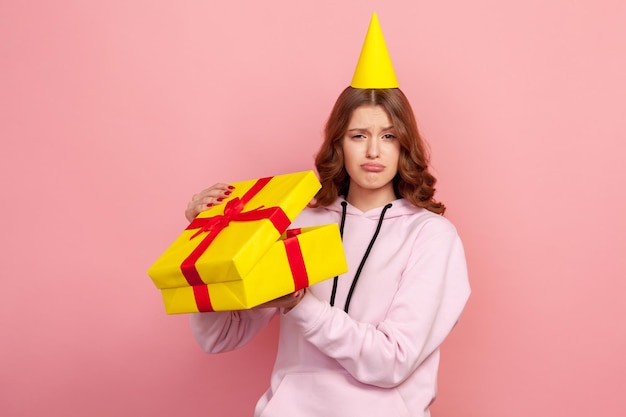 Porträt einer unzufriedenen jungen Frau in Hoodie und Partyhut, die ein Geburtstagsgeschenk auf einer Feiertagsfeier öffnet und enttäuscht aussieht. Studioaufnahme im Innenbereich isoliert auf rosa Hintergrund