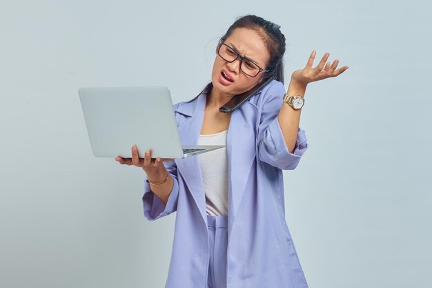 Porträt einer verärgerten jungen asiatischen Frau, die am Handy spricht und einen Laptop hält und zeigt, um Platz mit Palmen zu kopieren, die auf weißem Hintergrund isoliert sind