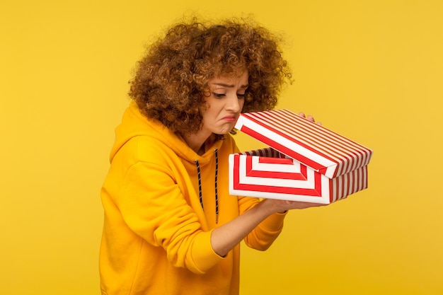 Porträt einer verärgerten, unzufriedenen, lockigen Frau in Hoodie, die in die Geschenkbox schaut, die das Geschenk öffnet, und mit unzufriedenem Ausdruck hineinspäht, Studioaufnahme isoliert auf gelbem Hintergrund