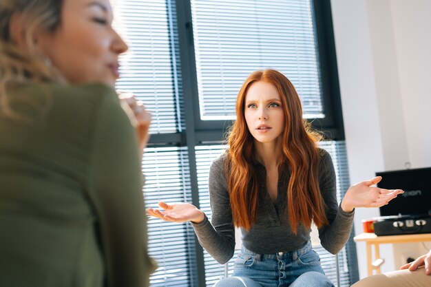 Porträt einer verblüfften jungen rothaarigen frau schaut weg mit den händen, die während des brainstormings von startprojekten in einem modernen büroraum in der nähe des fensters unverständnisvoll ausgebreitet sind.