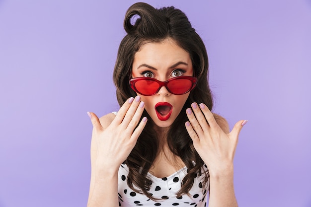 Porträt einer verführerischen Pin-up-Frau mit Retro-Sonnenbrille, die Wunder mit offenem Mund über violetter Wand isoliert ausdrückt